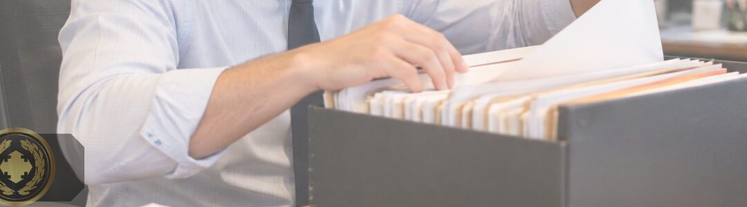 Funcionário de cartório procura um documento em uma caixa de arquivo sobre a mesa, em ambiente de escritório organizado e com clima institucional.