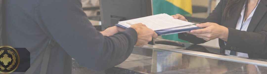 Pessoa estrangeira entrega documentos no atendimento de um consulado brasileiro, em um ambiente institucional, com a bandeira do Brasil ao fundo.